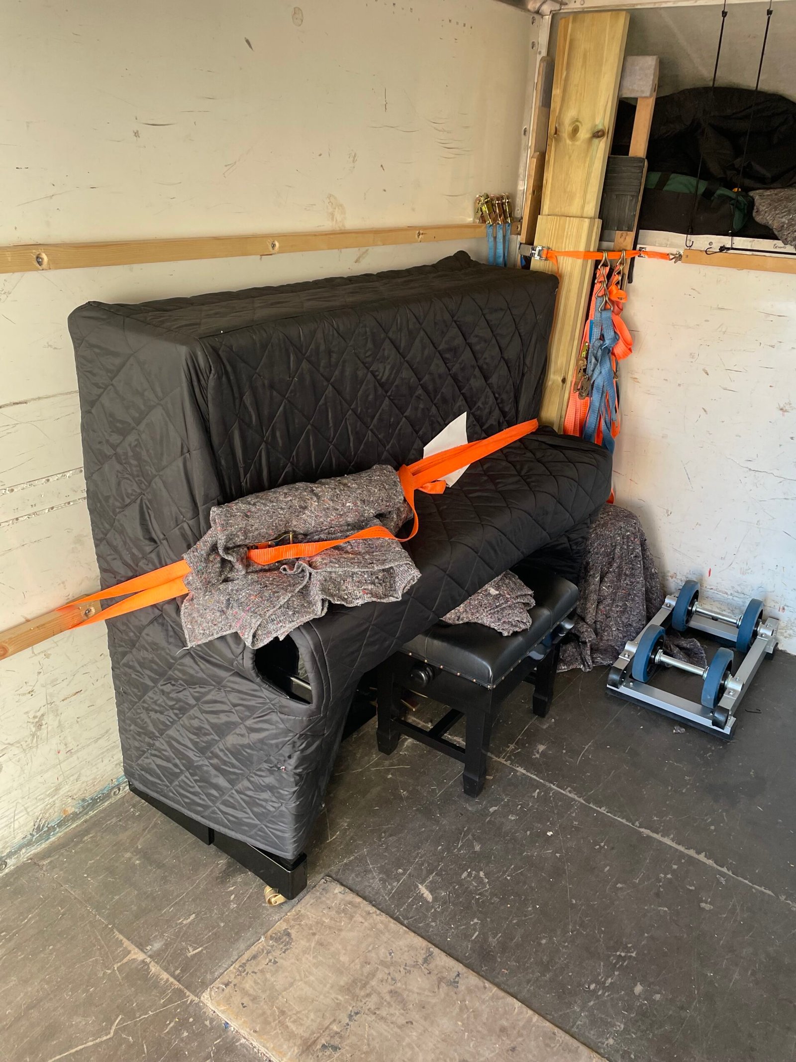 Upright piano wrapped in protective blanket and secured with an orange ratchet strap.