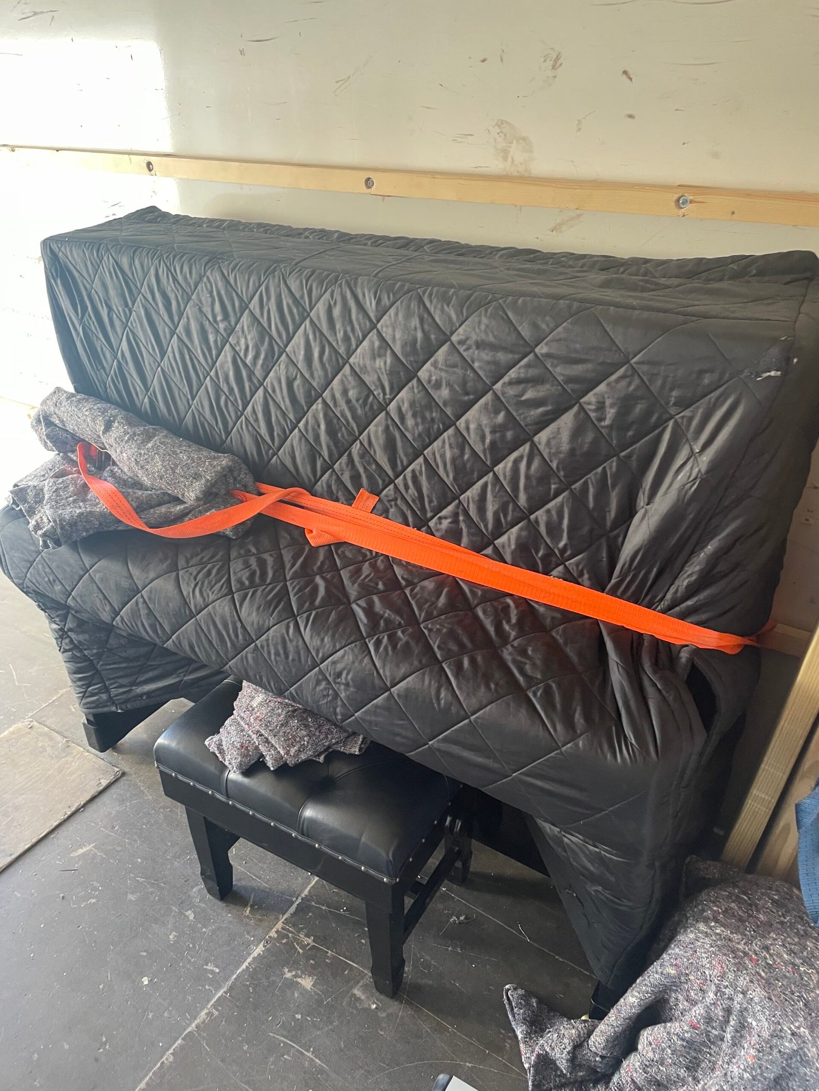 Upright piano wrapped in protective blanket and secured with an orange ratchet strap.